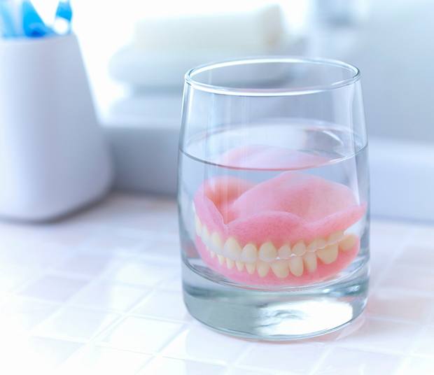 Set of dentures in a glass of water on a white tile bathroom countertop